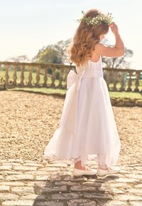 Ragazza in un abito bianco con un fiocco e una corona floreale si trova su un sentiero di pietra vicino a un balaustre di giardino sotto la luce del sole.