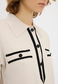 Blusa color crema con colletto a punta, dettagli con bottoni neri e strisce nere a contrasto su tasche e patta. Tessuto morbido.