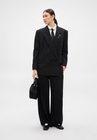 Μαύρο σακάκι διπλό με πλατιά πέτα, συνδυασμένο με φαρδιά μαύρη παντελόνα. Αξεσουάρ περιλαμβάνουν μια μαύρη τσάντα με υφή και μια γραβάτα.