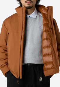 Mann in rostfarbener Steppjacke, die geöffnet ist, um den grauen The North Face Sweatshirt und das weiße Hemd darunter zu zeigen.