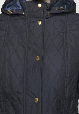 Gros plan sur un manteau matelassé bleu marine foncé à capuche, avec des boutons-pression dorés et des bloque-cordons.