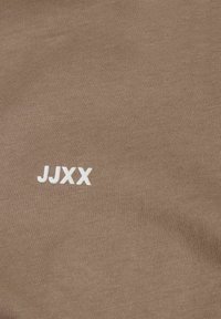 Tissu marron avec un petit logo blanc "JJXX" imprimé près du centre, montrant un gros plan de la texture du textile.