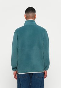 Mann iført en teal fleecejakke og blå jeans, vendt bort fra en ensfarget hvit bakgrunn.