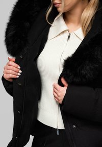 Černý zimní kabát s kapucí z umělé kožešiny, se zipovým zapínáním, v kombinaci s bílým žebrovaným svetr. Hladké textury vizuálně kontrastují.