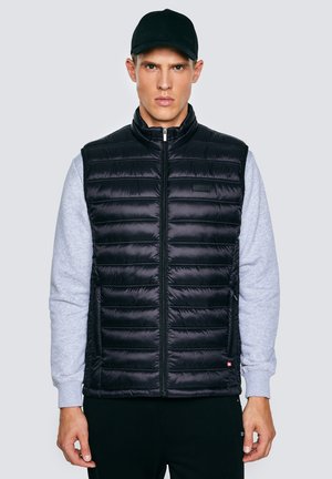 Noor mees musta tikitud vestiga halli pusa ja musta nokamütsiga, seisab heledusega ühevärvilise tausta ees.