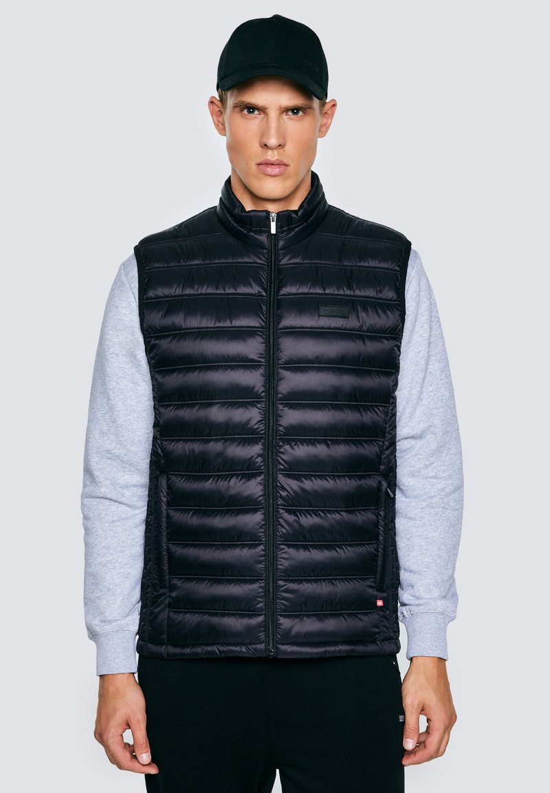 Jonge man die een zwart gewatteerd vest draagt over een grijze trui en een zwarte pet, staand tegen een effen lichte achtergrond.