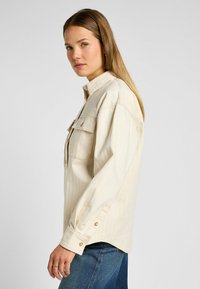 Camicia beige con bottoni, due tasche sul petto, maniche lunghe e taglio dritto. Presenta un colletto classico e dettagli con bottoni a scatto.