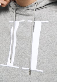 Sudadera con capucha de color gris claro, hecha de tela suave, que presenta un diseño impreso en blanco prominente y cordones grises con puntas de metal.