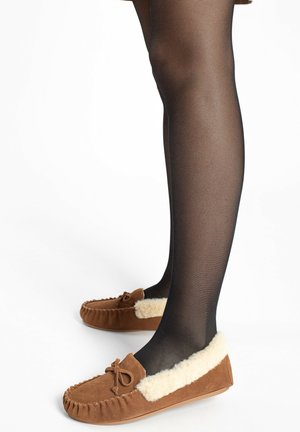 Bruine suedebotjes met crèmekleurige bontvoering, strikaccent en moccasinstiksel, gedragen met sheer zwarte panty’s. Eenvoudig en laag profiel ontwerp.