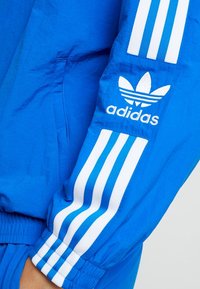 Blå Adidas-jacka med vita ränder längs ärmarna. Innehåller Adidas-logotypen på överarmen. Materialet verkar lätt och slätt.