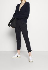 Marinblå oversized tröja, beige topp, svarta tapered byxor, vita sneakers med svarta accenter, håller en svart kviltad väska, avslappnad outfit.