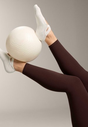Ben i mørkebrune leggins og hvide sokker holder en struktureret hvid træningsbold mellem hævede fødder mod en neutral baggrund.