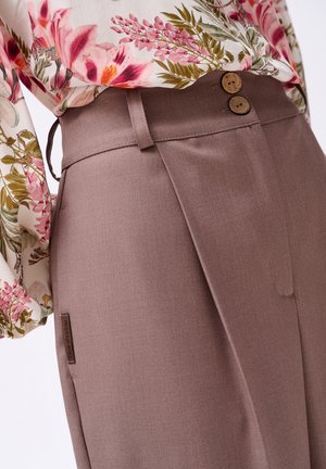 Braune maßgeschneiderte Hose mit glatter Textur, ausgestattet mit zwei dekorativen Holzknöpfen und einem Seitenschild. Kombiniert mit einer blumenmusternden Bluse.