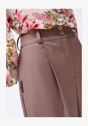 Braune maßgeschneiderte Hose mit glatter Textur, ausgestattet mit zwei dekorativen Holzknöpfen und einem Seitenschild. Kombiniert mit einer blumenmusternden Bluse.