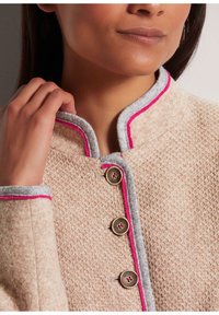 Texturierte beige Jacke mit grauem Kragen, rosa Paspel und dekorativen braunen Knöpfen. Nahaufnahme des Kragens und der oberen Frontdetails.