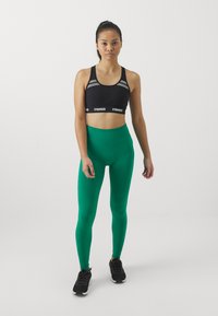 Zwarte sportbeha met grijze accenten, gecombineerd met hoogtaille groene leggings. Model draagt zwarte sportschoenen. Eenvoudig en aansluitend ontwerp.