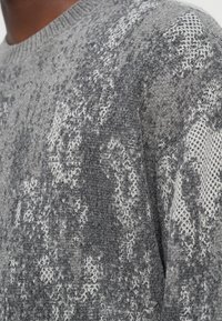 Γκρι πλεκτό πουλόβερ με υφή και διάφορες αποχρώσεις του γκρι και του λευκού, στρογγυλή λαιμόκοψη και μαλακό ύφασμα.