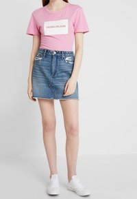 Rosa Baumwoll-T-Shirt mit weißem Rechteck-Logo, kombiniert mit einem blauen Jeansrock mit ausgefranstem Saum und verwaschenen Details. Weiße Sneakers.