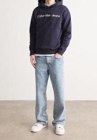 Marinblå hoodie med vit "Calvin Klein Jeans"-logotyp, kombinerad med ljusblå urblekta denimjeans och vita sneakers.