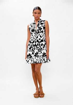 Mujer con vestido sin mangas de flores en blanco y negro y sandalias marrones, de pie contra un fondo blanco liso.