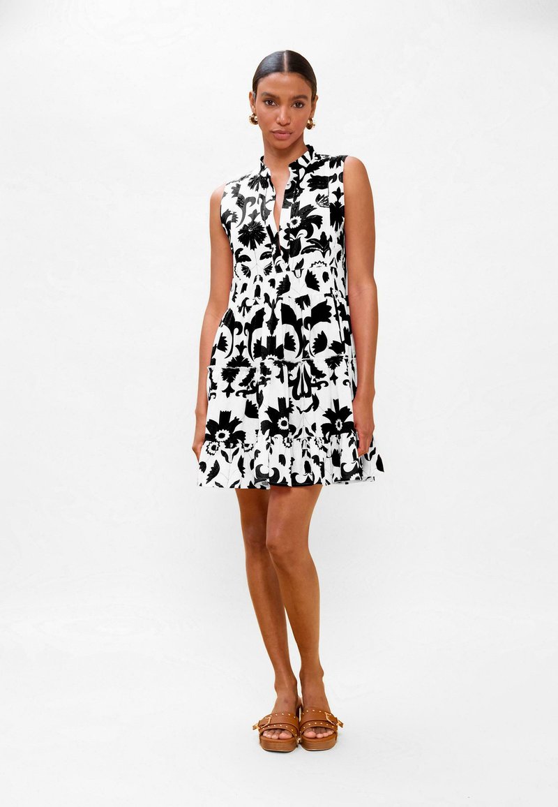 Femme en robe sans manches à fleurs noires et blanches et sandales marron, debout devant un fond blanc uni.