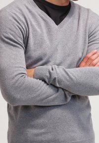 Pull gris en maille avec un col en V. Présente des poignets et un ourlet côtelés, une texture douce et une coupe ajustée. Porté sur un haut noir.