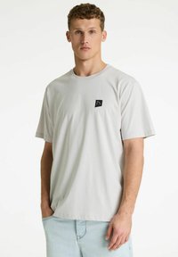Joven de piel clara y cabello rizado que lleva una camiseta gris claro lisa con un pequeño logo negro en el pecho y unos vaqueros azul claro.