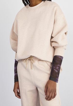 Person iført beige oversized sweatshirt og matchende sweatpants med sorte og lilla tie-dye lange ærmer synlige under.