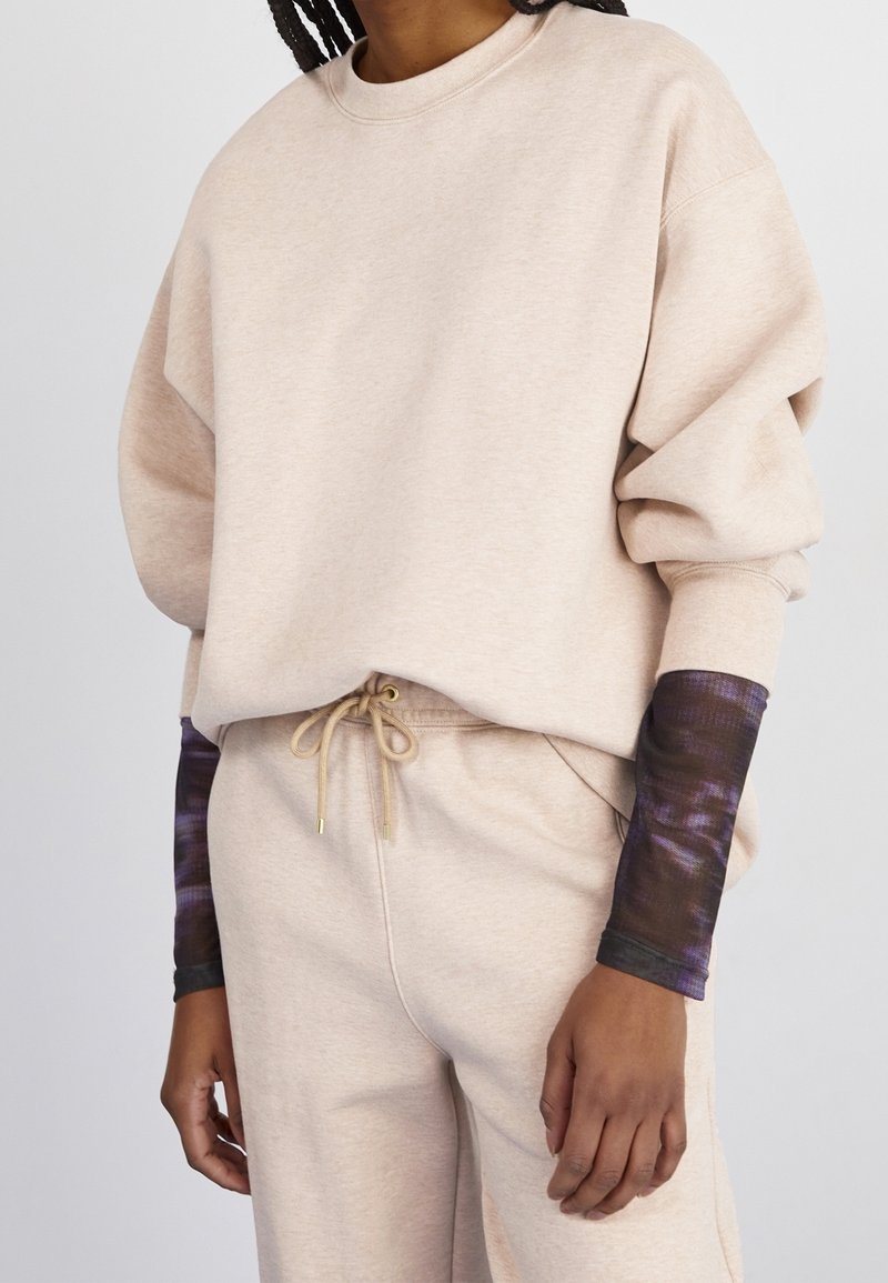 Personne portant un sweat-shirt beige oversize et un pantalon de jogging assorti avec des manches longues tie-dye noires et violettes visibles en dessous.