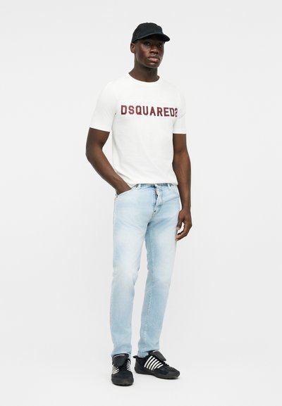 Man wearing black cap, white DSQUARED2 t-shirt, light blue jeans, and black sneakers with white stripes, standing with one hand in pocket.