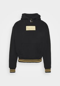 Sudadera negra con un parche de logo bordado en oro, bolsillo tipo canguro en la parte frontal, y puños y dobladillo acanalados con franjas doradas y amarillas.