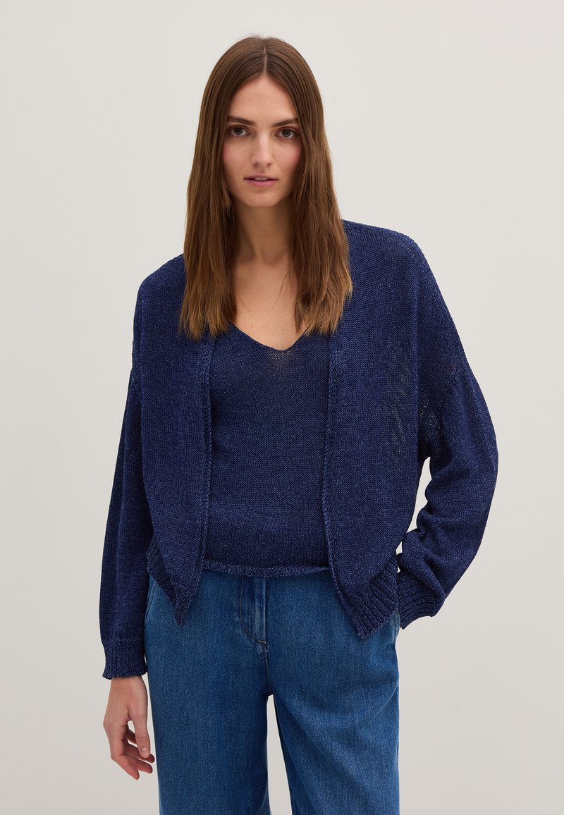 Marineblaue Strickjacke mit verlängerten Ärmeln und offenem Front, kombiniert mit einem passenden V-Ausschnitt-Oberteil. Getragen mit hochgeschnittenen Jeanshosen.