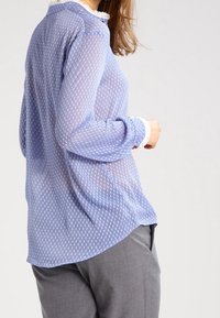 Blouse bleu clair translucide à pois blancs, manches longues et col à volants, associée à un pantalon gris.