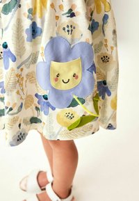 Niña vestida con un vestido de estampado floral que tiene un gran aplique de flor azul sonriente y sandalias blancas sobre un fondo claro.