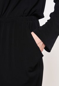 Černý oděv s volným střihem, elastickým pasem, dlouhými rukávy a bočními kapsami. Látka vypadá hladce a lehce.