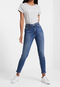 Kobieta w szarej koszulce Calvin Klein Jeans, w niebieskich wysokich dżinsach skinny i białych trampkach, stojąca w rozkroku.