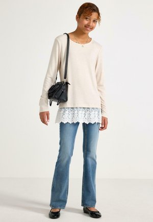Ung kvinde smiler, iført beige langærmet top med blonderkant, blå jeans, sorte ballerinaer og bærer en sort skuldertaske.