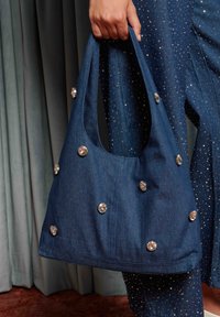 Džinsa tote soma ar izliekta dizainu. Aprīkota ar apaļu rokturi, izkliedētām kristāla pērlītēm un tumši zilu teksturētu audumu.