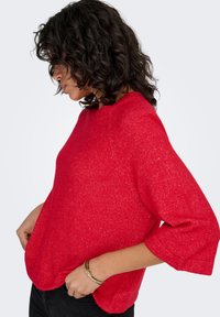 Pull rouge texturé avec un col rond et des manches trois-quarts, doté d'une coupe ample et d'une légère variation à l'ourlet.