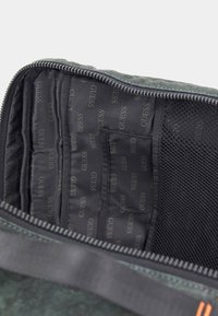Zelené textúrované vnútro tašky s čiernou podšívkou, ktorého súčasťou je opakujúce sa logo "GUESS". Obsahuje sieťovanú kapsu a viacero slotov na karty. S otvorom na zips.