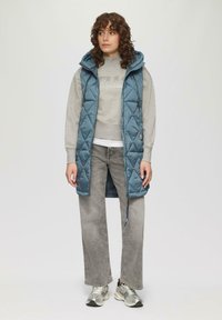 Warpierte teal Weste mit quadratischem Design, Kapuze und Seitenfächern, kombiniert mit einem grauen Sweatshirt und grauen weit geschnittenen Jeans sowie metallischen Sneakers.