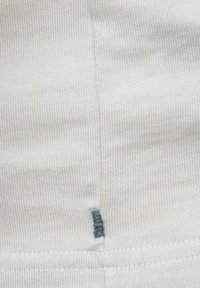 Tessuto bianco lavorato a maglia con una texture a coste e un dettaglio di cucitura visibile. È presente un piccolo punto blu vicino alla cucitura centrale.