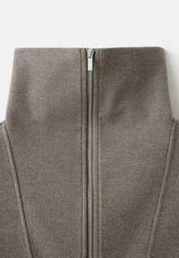 Siva jopa z zadrgo in visokim ovratnikom, narejena iz mehkega materiala. Ima srebrno zadrgo na sredini in šive na straneh.