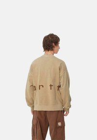 Sudadera beige con un corte relajado y hombros caídos, con el texto marrón "earth" en la parte posterior. Combinada con pantalones cargo marrones.