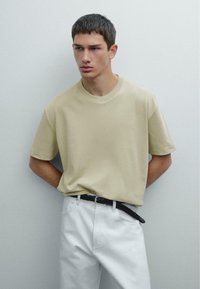 Beige tričko s krátkým rukávem z měkké tkaniny, kulatý výstřih, v kombinaci s bílými kalhotami a černým pleteným páskem. Jednoduchý a moderní design.