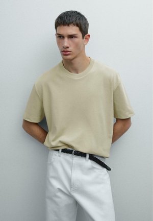 Beige short-sleeve t-shirt in soft fabric, round neckline, paired with white pants and a black braided belt. Simple and modern design.