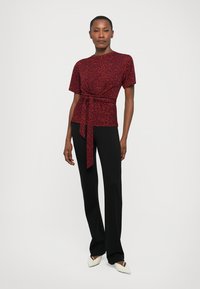 Vrouw die staat in een rode, gepatteerde top met korte mouwen en een tailleband, zwarte broek en wit, puntig geschoeide schoenen tegen een effen achtergrond.