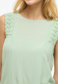 Miętowa zielona bluzka z koronkową lamówką na ramionach, gładką teksturą, okrągłym dekoltem i subtelnym marszczeniem po bokach.