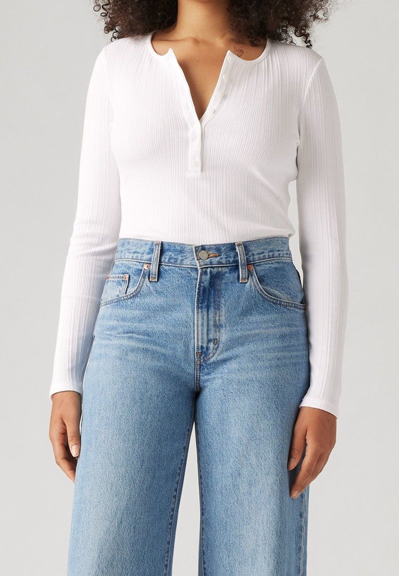 Frau trägt ein weißes, geripptes Langarm-Henley-Shirt, das in eine hochtaillierte hellblaue Jeans gesteckt ist, und steht vor einem schlichten Hintergrund.