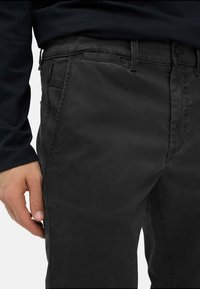 Sorte bomulds chinos med en glat tekstur, der har sidelommer og en streamlined pasform. Nærbillede viser stofdetaljer og syninger.